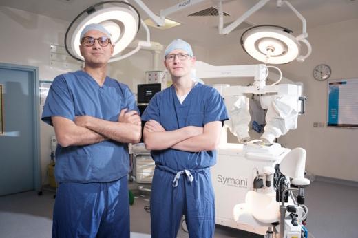 Two men in blue scrubs, in front of a surgical robot