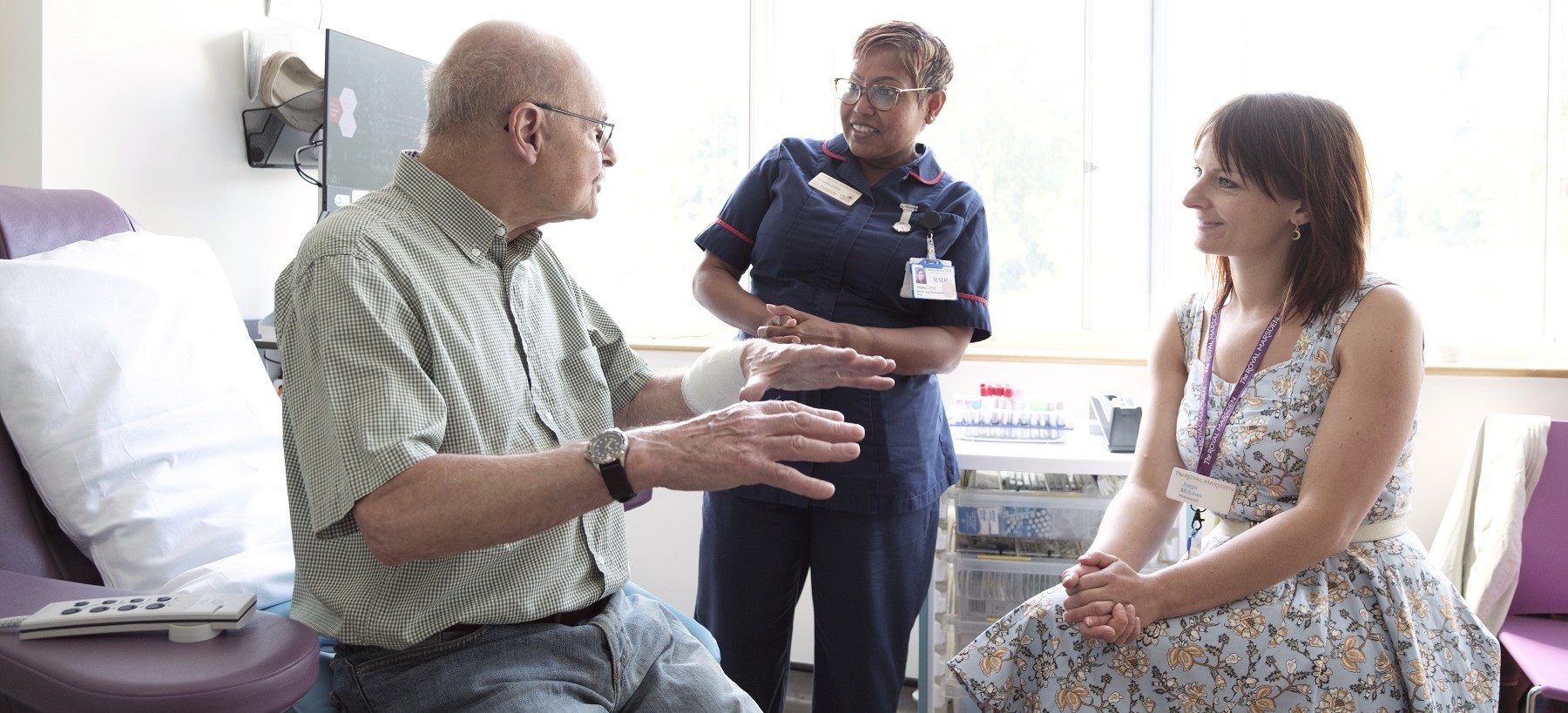 A patient sitting in a hospital room talking to two healthcare professionals who are smiling and listening to them intently