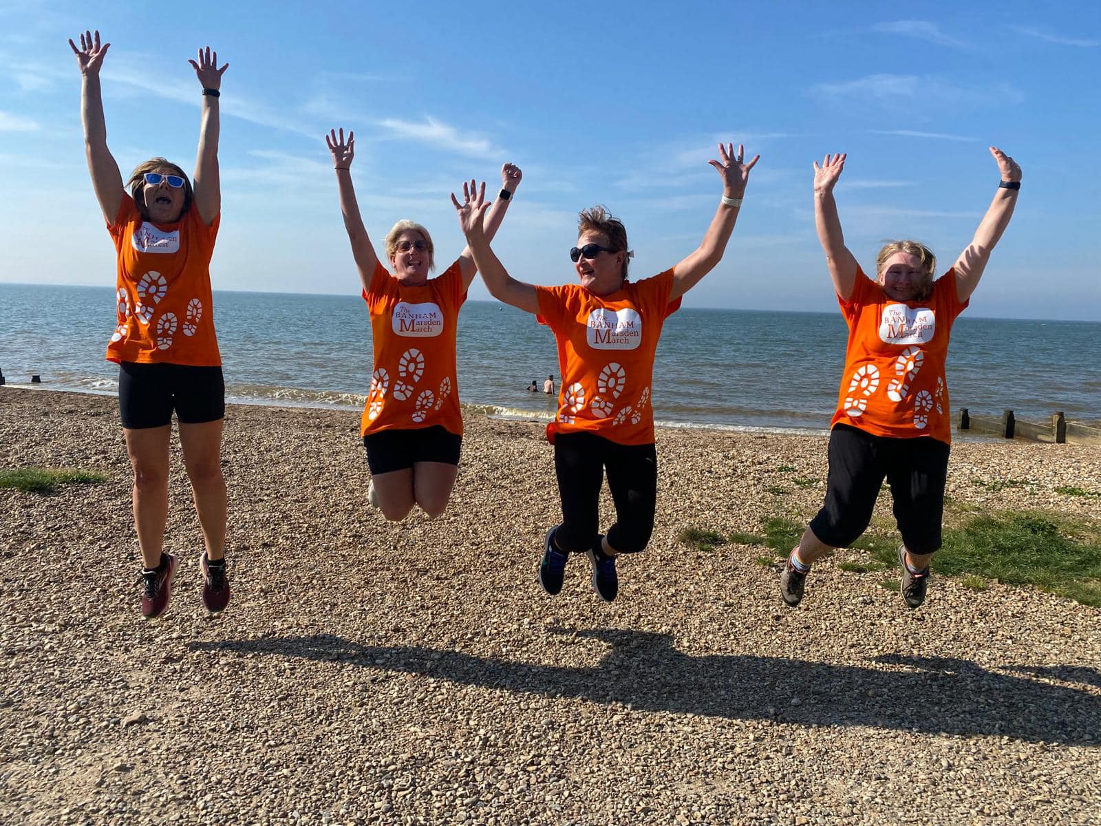 Four women wearing orange Banham Marsden March t-shirts jumping together on a beach.