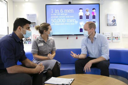 A dark-haired man sits next to a woman in a nurse's uniform, they are talking to HRH The Duke of Cambridge who is sat opposite them.  There is a digital screen behind them, all are wearing surgical masks