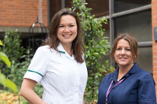 Two women stood outside smiling. The woman on the left is tall and wearing a white uniform. The woman on the left is shorter and wearing a navy blue nurses uniform.