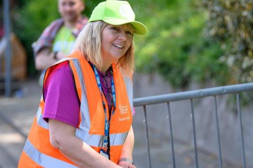 A woman smiling. She is wearing a Banham Marsden March volunteer vest and cap and by a table of finishers medals. 