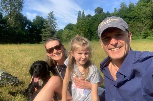 A family of three and their small black dog smiling and sitting in a grassy field in the sunshine