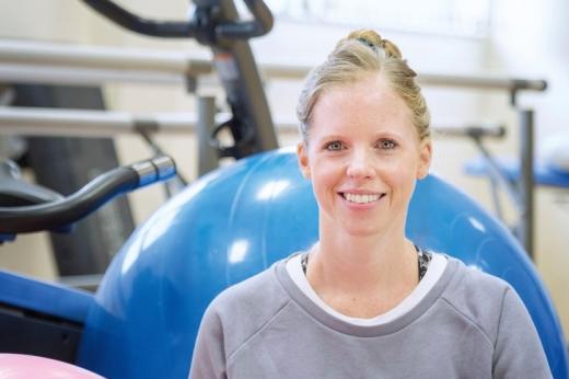 A Royal Marsden Physiotherapist smiling, wearing a sports jumper and sitting in front of gym equipment including a yoga ball and exercise bike.
