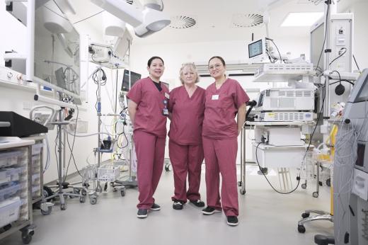 Three people in pink hospital scrubs, standing in a hospital room filled with medical equipment