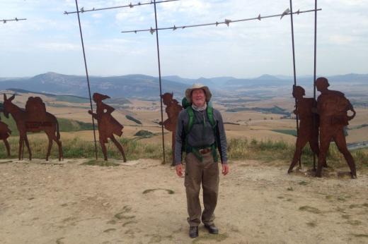 A Man stands at a view point with hills in the background. He is wearing hiking gear and a floppy tan hat.