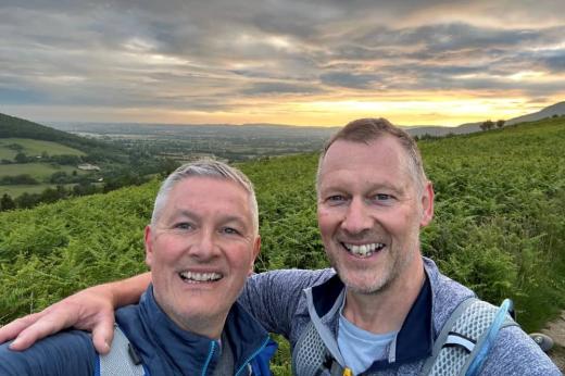 A selfie of two smiling men with a sunset and moorland in the background. They have their arms round each others shoulders. 