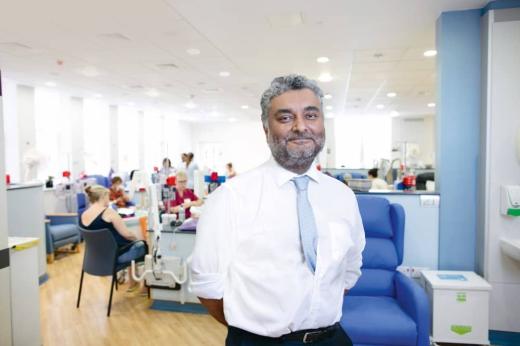 A smiling man in a white shirt in a hospital unit. He has his hands behind his back and a light blue tie on. 