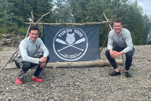 Hector and Charlie outdoors, crouching down in front of a Yukon 1000 banner