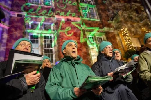 A group of men in blue wooly hats stand infront of the lit up facade of The Royal Marsden hospital in Chelsea. They are singing carols.