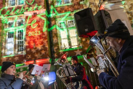 A wide angle shot of a brass band playing carols. They are playing bright silver instruments. In the background a red brick building is lit up with stars and lights. 