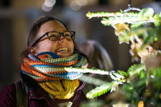 A woman in a stripy knitted scarf and black glasses smiling up at the Christmas tree