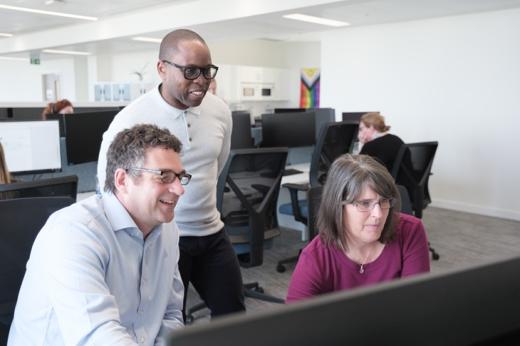 Three people in an office around a computer. One man is standing, and a man and woman are seated.