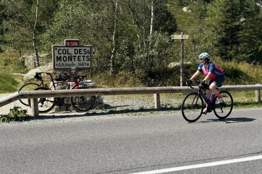 Jenni cycling on a road. There is a lot of green foliage and trees in the background, with a sign that reads 'Col Des Montets, Altitude 1461m'