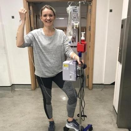 A woman smiling and standing in a Royal Marsden hospital corridor hooked up to a drip on wheels, through a cannula in her arm