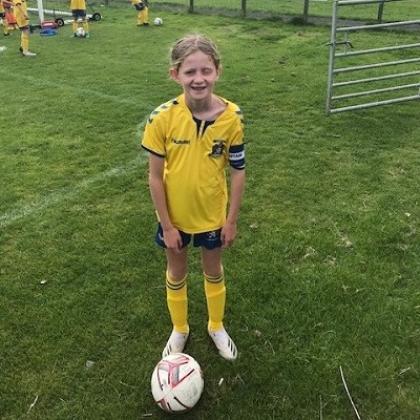 A young girl standing and smiling on a football pitch, wearing a bright yellow football kit with a ball by her feet