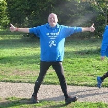 A man walking through a park, smiling with his arms out wide and giving two big thumbs up. He's wearing a bright blue hoody that reads '5k your way'