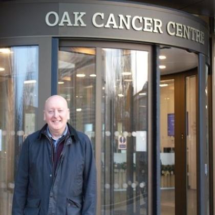 A man in a jacket and coat, standing outside the entrance to the Oak Cancer Centre