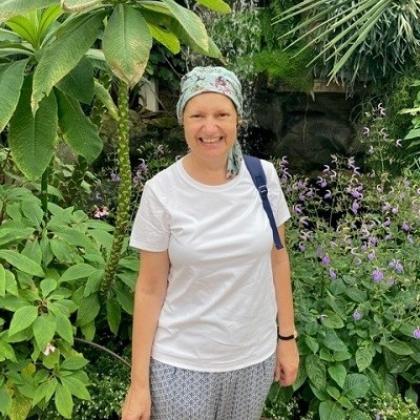 A woman wearing a summery outfit and a headscarf, standing and smiling in front of some tropical plants