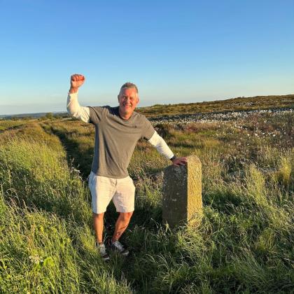 Paul standing on the moors at sunset with his fist in the air. He has his other hand on a route marker stone. 