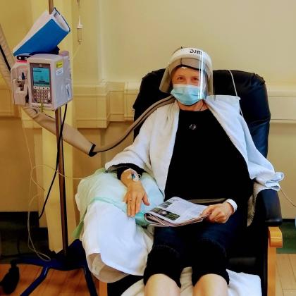 A woman sat in a treatment chair holding a magazine. She has a towel around her shoulders and is wearing a blue medical mask