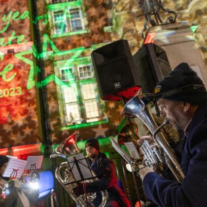 A wide angle shot of a brass band playing carols. They are playing bright silver instruments. In the background a red brick building is lit up with stars and lights. 