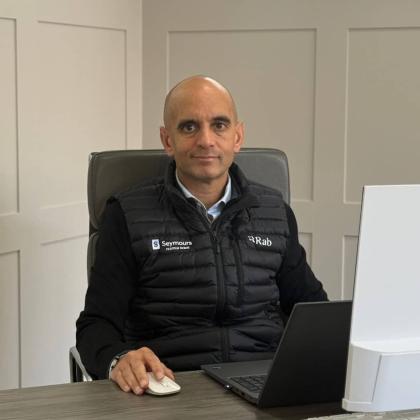 A bald man in a black jacket sat at a desk computer. He is smiling to the camera.