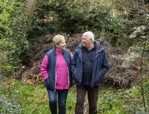 An older couple  smiling and looking at each other as they walk arm in arm through woodland.