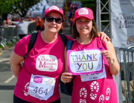 Two smiling brunette women in pink Banham Marsden March t-shirts and caps. They are holding up a hand drawn sign saying 'Thank you'