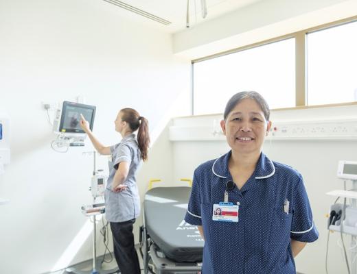 A nurse wearing a blue uniform and name badge smiling and standing in a bright, airy medical room in the Oak Cancer Centre. With a another nurse working in the background.