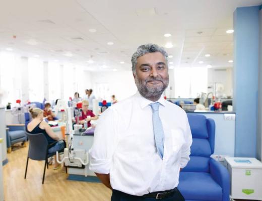 A smiling man in a white shirt in a hospital unit. He has his hands behind his back and a light blue tie on. 