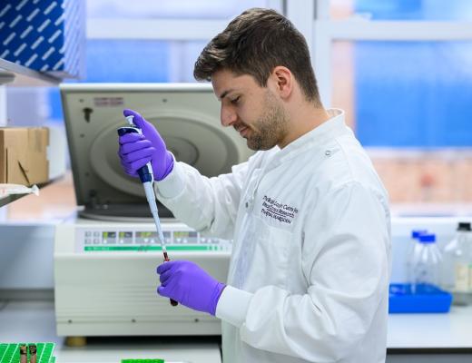 A scientist in a laboratory at The Royal Marsden. He is wearing a lab coat with rubber gloves, and is squeezing a substance into a test tube using a pipet.