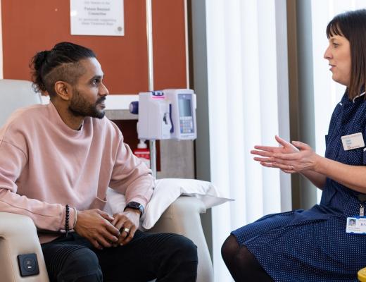 Royal Marsden patient, Sam, sitting on a comfy treatment armchair, listening to a nurse who is sitting and talking to him. She is wearing a blue uniform, with a Royal Marsden badge.