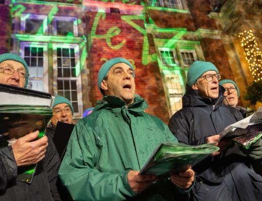 A group of men singing carols outside a lit up building. They are wearing matching teal blue woolly hats. 