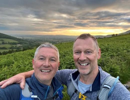 Two men smiling in front of mountains and a sunset