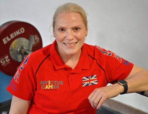 Amanda smiling and wearing a bright red Invictus Games UK shirt. She's leaning on a large weight in a gym. 