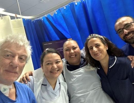 Mike, with a tracheostomy tube in his neck standing with a group of smiling staff at The Royal Marsden. They are all wearing scrubs and standing in a hospital room. 