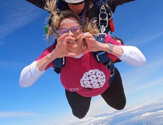 Woman in pink RMCC t-shirt making a heart with hands mid-skydive