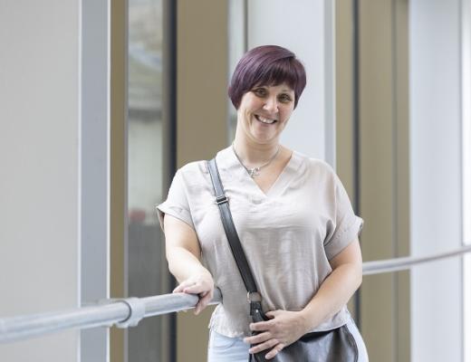 Roxanne, bowel cancer patient at The Royal Marsden, standing and smiling in front of the bright window of the Oak Cancer Centre.. She has short, purple/brown coloured hair, and is wearing a light top with a black handbag over her shoulder.