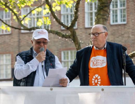 Two men speaking at the startline podium. The man on the left is Charles Hallat, the CEO of Banham. He is wearing a shirt and jacket, and a white 'Team Banham' Banham Marsden March cap. 