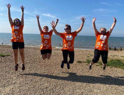 Four women wearing orange Banham Marsden March t-shirts jumping together on a beach.