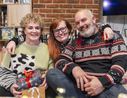 Three people smiling with their arms round each other. Zoe, on the left, is wearing a green jumper and holding christmas presents, Chloe in the middle is wearing a patterned Christmas jumper. She has long auburn hair and is wearing glasses. Marc is on the right, he has a white beard and is wearing a Christmas jumper with reindeer on it.  