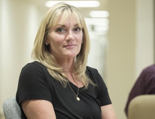 Upper body shot of a woman sitting, looking at the camera. She has blonde hair and is wearing a black top