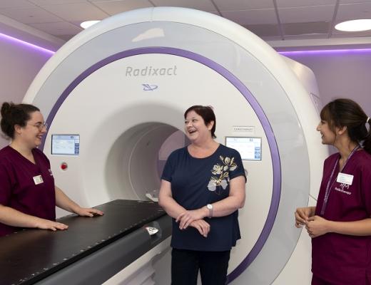 A patient smiling and laughing with two people from The Royal Marsden's radiotherapy team. They are standing in front of a large white Radixact machine, which has a lateral bed for patients to lie down and a tunnel, where patients' heads go.