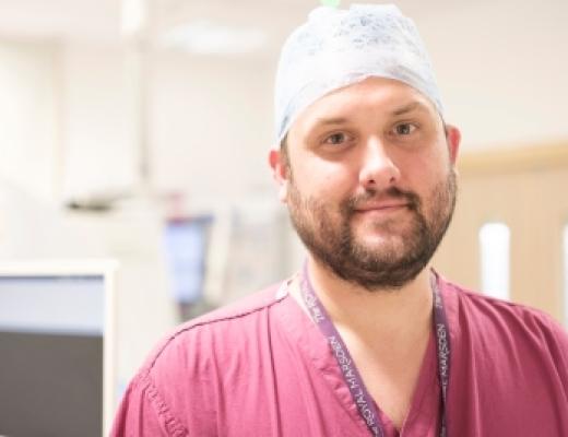 A man, Dr Ed Johnston, looks into the camera. he has a light brown beard and a blue scrub cap on. He is wearing pink scrubs and a Royal Marsden lanyard.