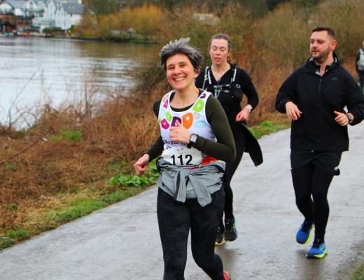 A Team Marsden runner smiling and running along a riverside path for the Palace Half Marathon.  is running on a path that runs alongside the river Thames.