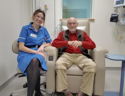 Patient, Richard, is smiling and sitting on a comfortable leather treatment chair in an empty, quiet medical room at The Royal Marsden. Sitting next to him in a swivel office chair is his nurse who is wearing a bright blue uniform and name badge.