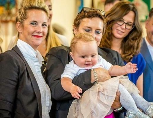 Two people dressed formally at a gathering of people. One of them is smiling at the camera, the other is holding a happy smiley baby, she is wearing a little ballet dress with tights and shiny trainers.