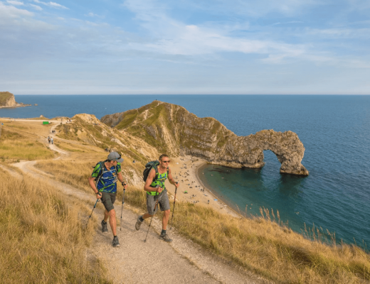 Two trekkers walking along a sandy coast line, next to blue sea and sky